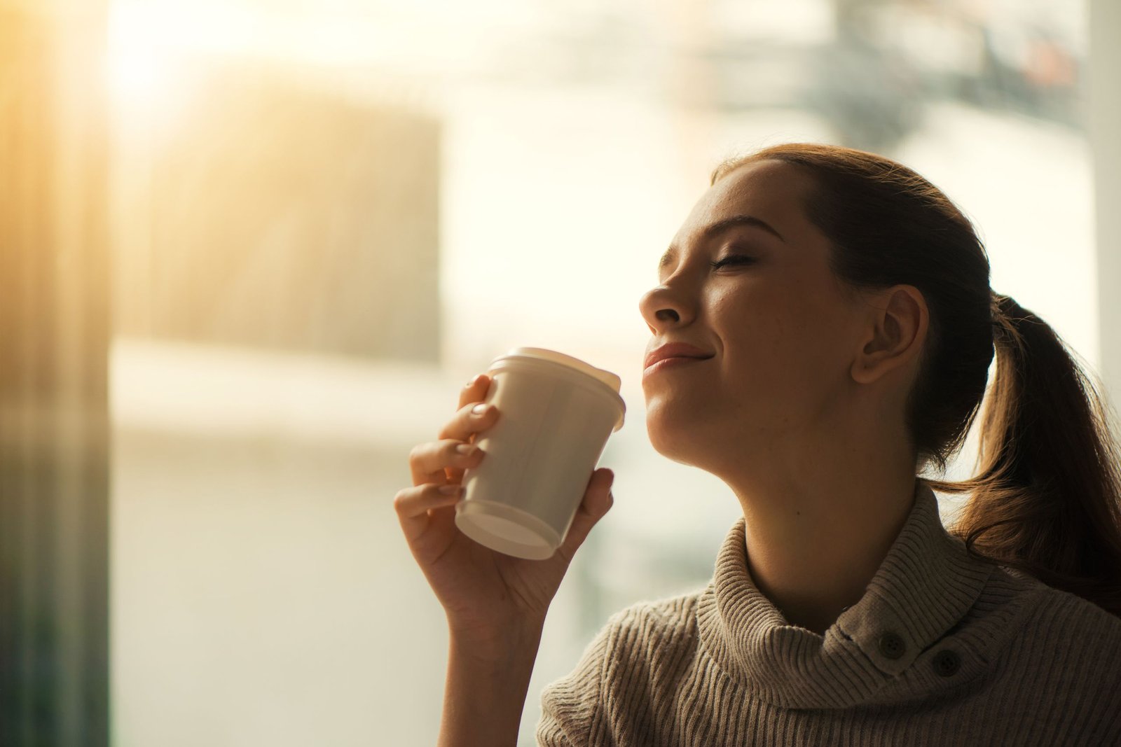 Smiling woman savoring morning coffee by the window in soft warm light.